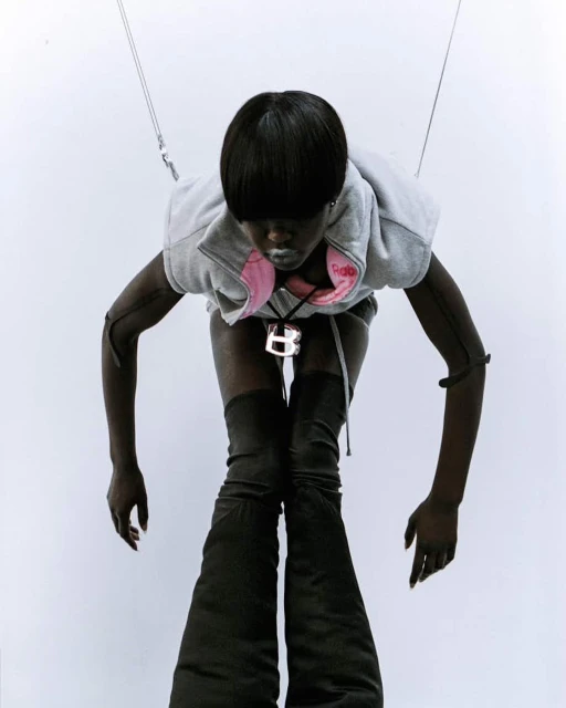 Woman with short black hair wears a grey hoodie vest and high, black, wide-legged boots against a white background in a fashion shot.