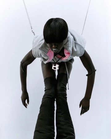 Woman with short black hair wears a grey hoodie vest and high, black, wide-legged boots against a white background in a fashion shot.