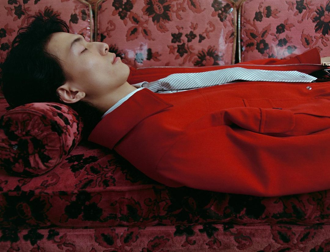 An Asian man lies on a floral couch wearing a red jacket in a medium shot. He has his eyes closed and head resting on a floral pillow.