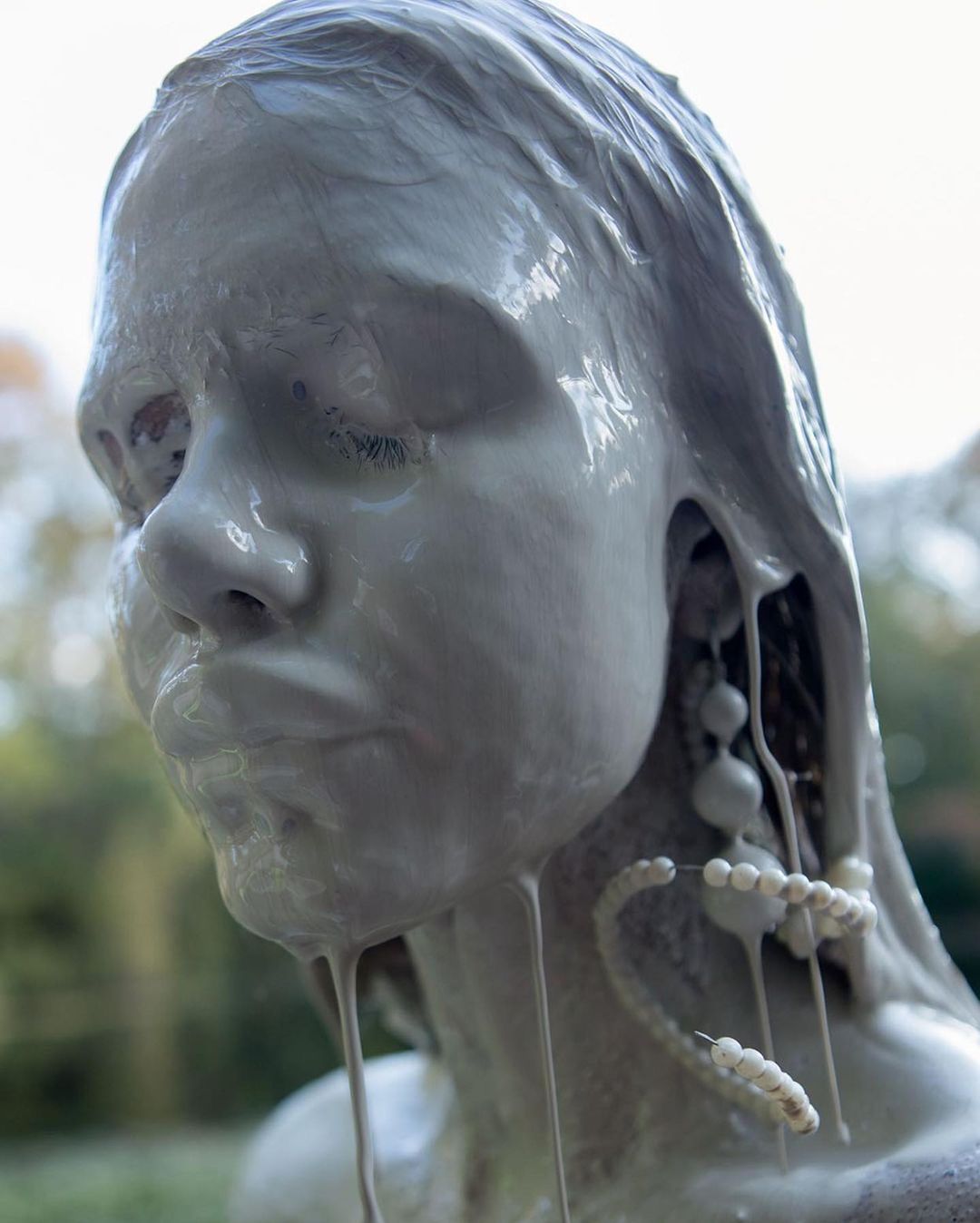 Close-up of a young person's face covered in light grey paint or clay, some of which is dripping, with beaded jewelry visible.