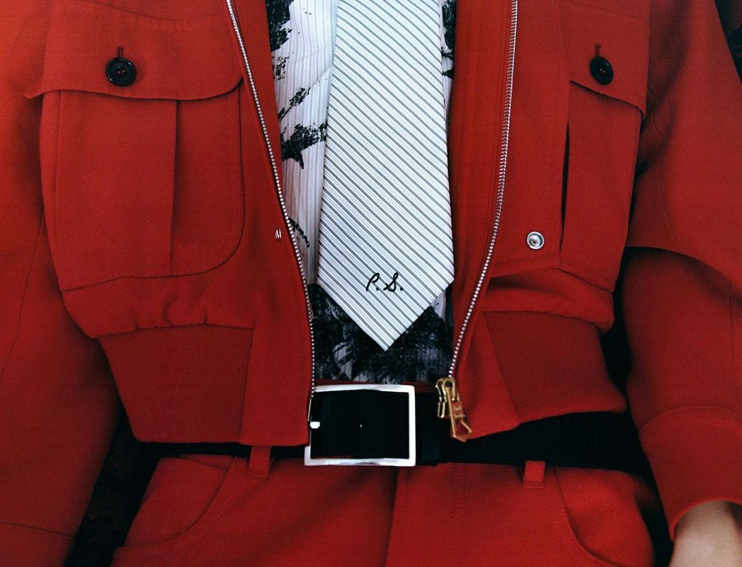Close up of a person wearing a red jumpsuit, a white and black shirt, and a striped tie, accessorized with a black belt with a silver buckle.