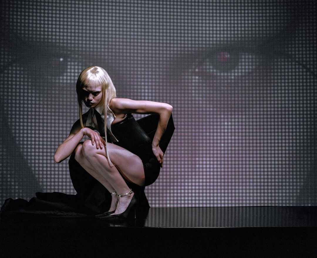 Blonde woman in dark fashion poses in front of a grayscale eye projection, creating a moody and dramatic visual narrative in a studio setting.