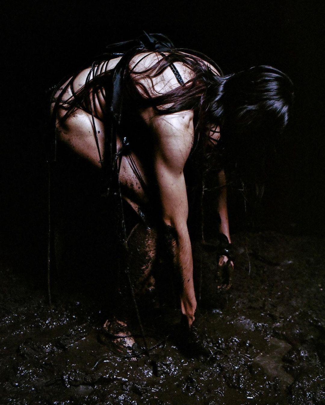 Woman stands knee-deep in muddy water, partially nude and covered in mud in a dimly lit, dark environment. Black ropes are around the body.