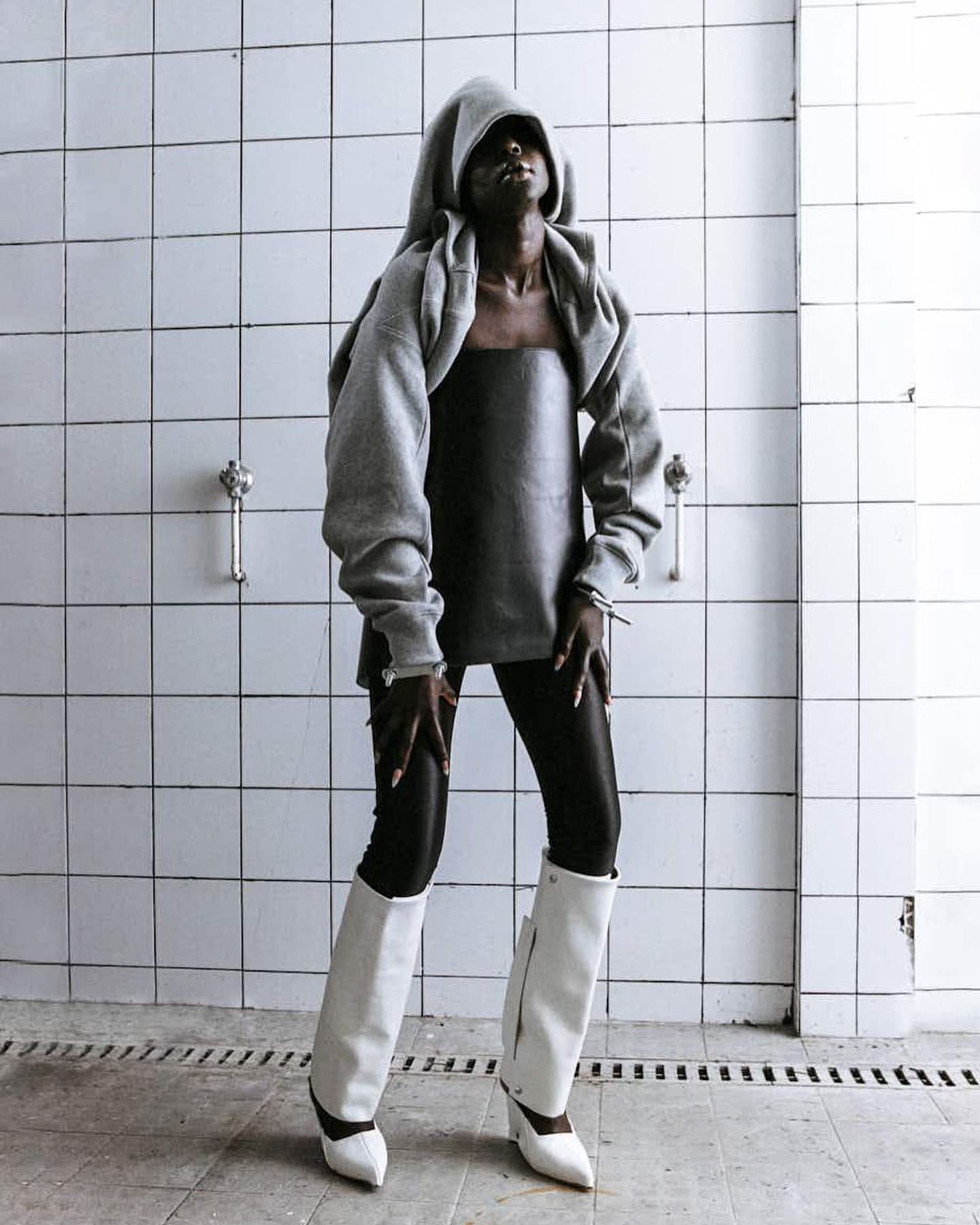 Woman in grey hooded sweater, black leggings and white boots strikes a pose against a tiled wall, looking upwards in an indoor setting.