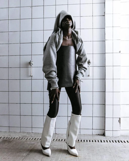 Woman in grey hooded sweater, black leggings and white boots strikes a pose against a tiled wall, looking upwards in an indoor setting.