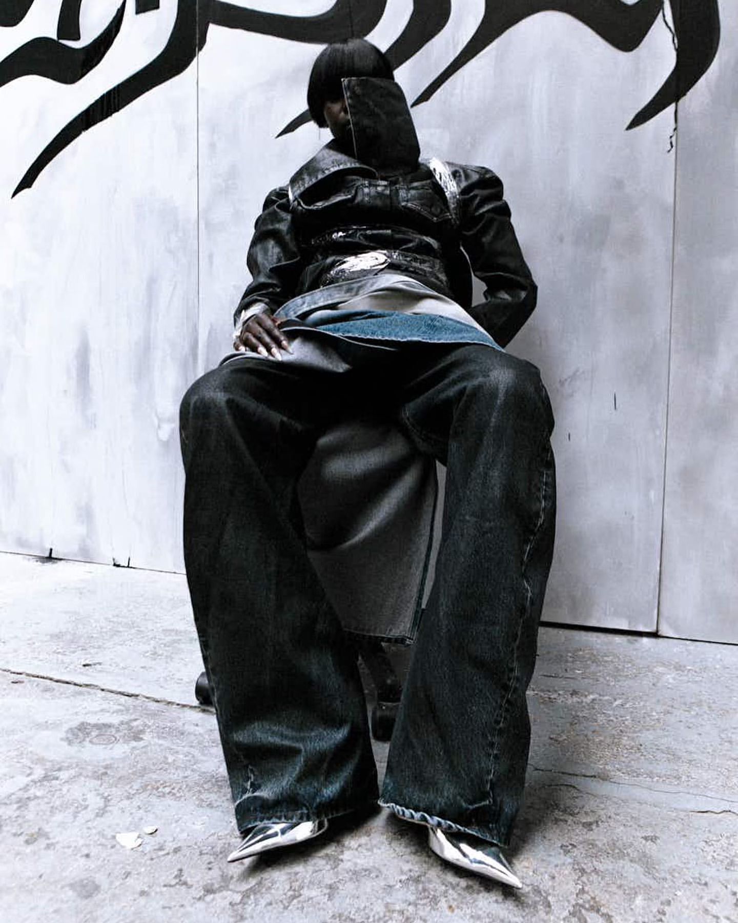 Fashionable person sits wearing avant-garde denim jacket, dark jeans, and shiny silver shoes against graffiti-style wall backdrop.
