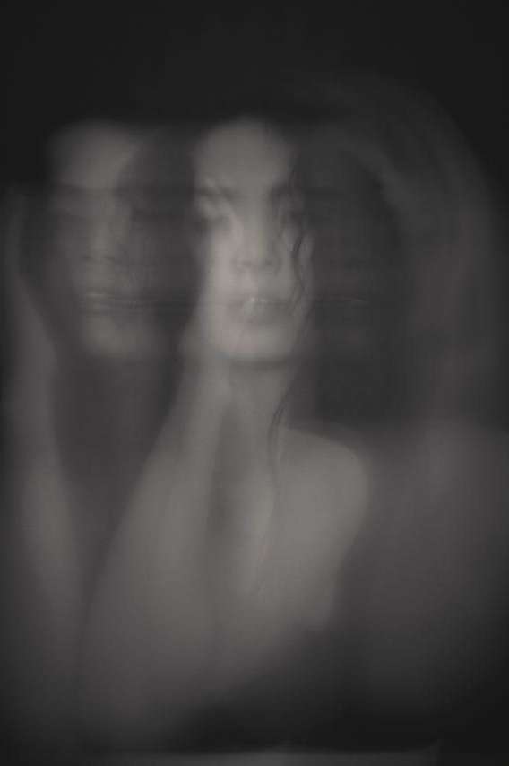 Monochromatic portrait of a young adult woman with a blurred, long-exposure effect and hands near her face against a dark backdrop.