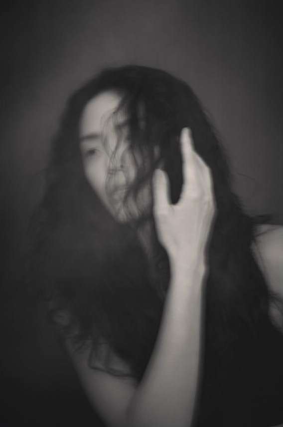 Monochromatic portrait of a blurry young woman with long dark hair and hand near her face on a dark grey seamless background.