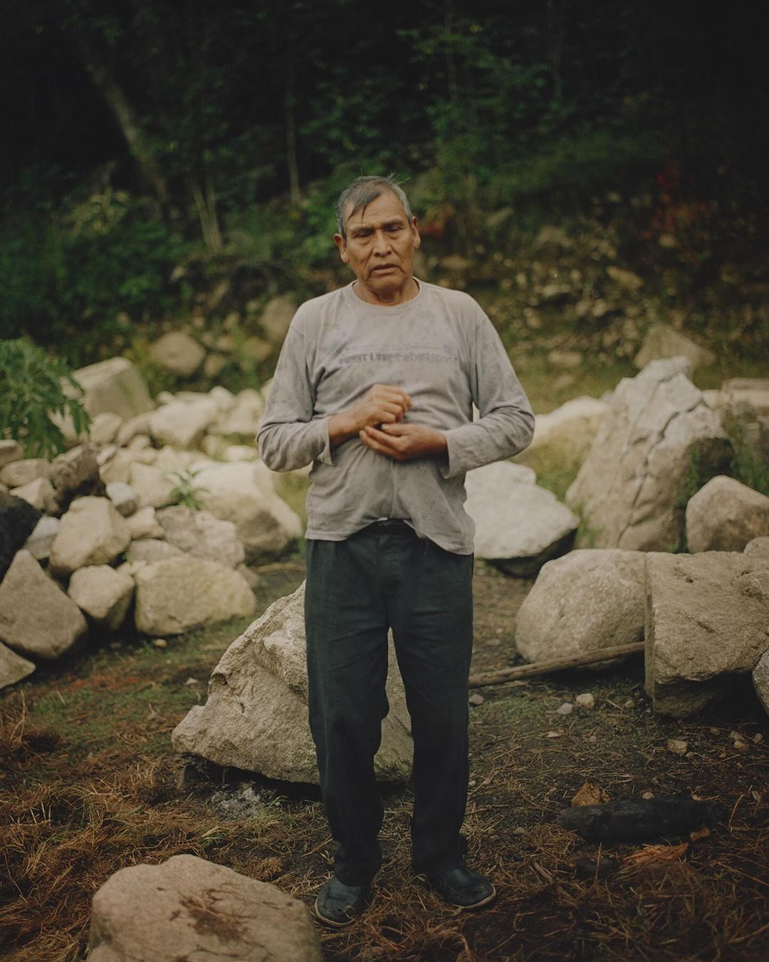Portrait of an adult male with hands clasped standing in a natural outdoor environment with rocks and vegetation in the background.