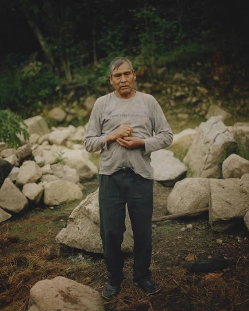 Portrait of an adult male with hands clasped standing in a natural outdoor environment with rocks and vegetation in the background.