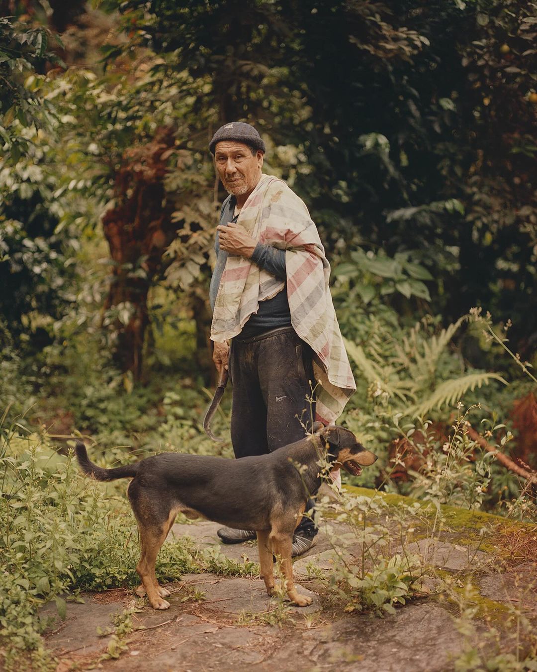 Elderly man stands outdoors with a dog in a verdant, natural setting, wearing a knit cap, dark pants, and a colorful shawl.