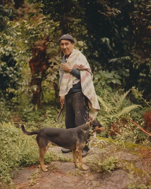 Elderly man stands outdoors with a dog in a verdant, natural setting, wearing a knit cap, dark pants, and a colorful shawl.