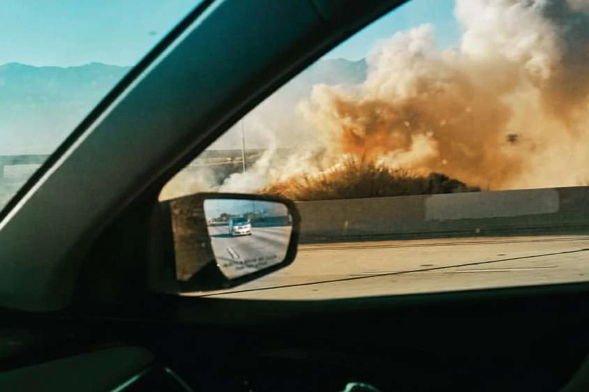 Car interior view of roadside fire and smoke plume near highway, seen through passenger window and side mirror of car.