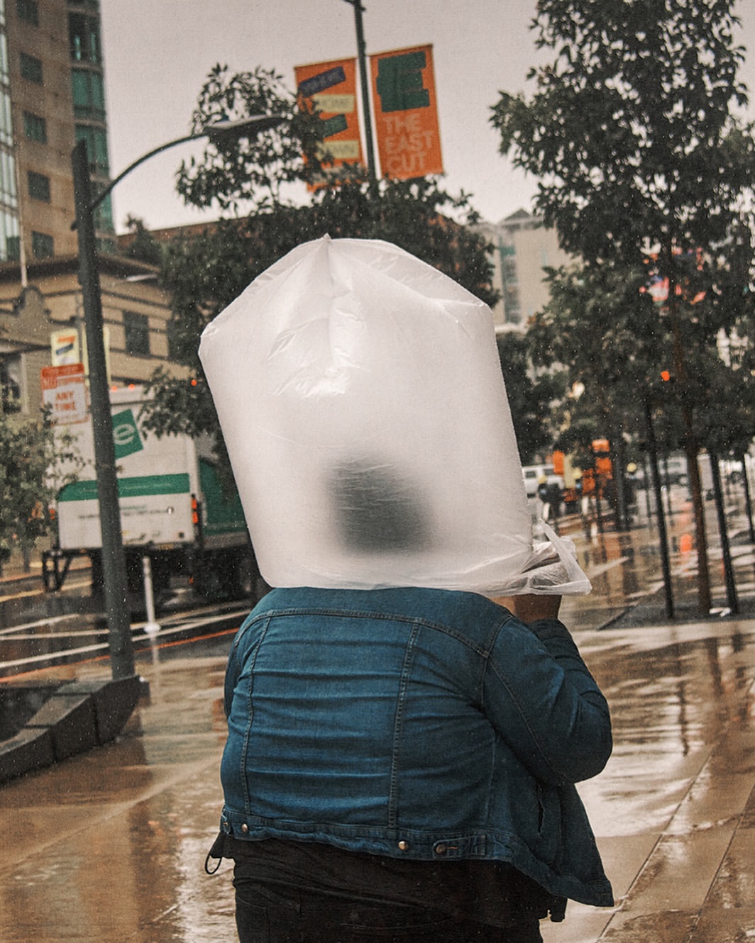 Person in a blue denim jacket walks in the rain-soaked city street with a transparent plastic bag over their head for rain protection.