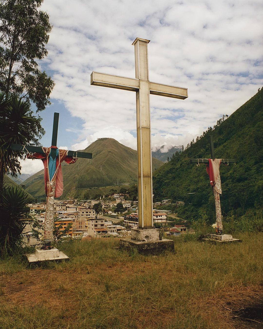 Three religious crosses stand on a grassy hill overlooking a town in a valley surrounded by forested mountains under a cloudy sky.