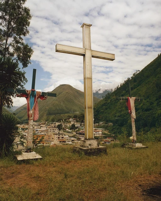 Three religious crosses stand on a grassy hill overlooking a town in a valley surrounded by forested mountains under a cloudy sky.