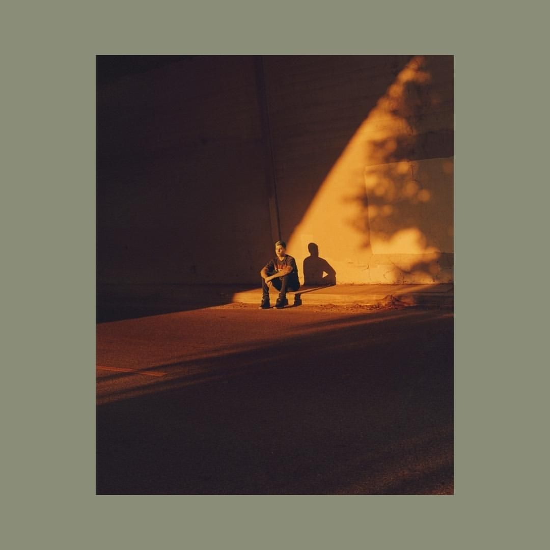 Adult male squats in pool of golden light with long shadow against neutral wall, wearing jeans, t-shirt, and sneakers.