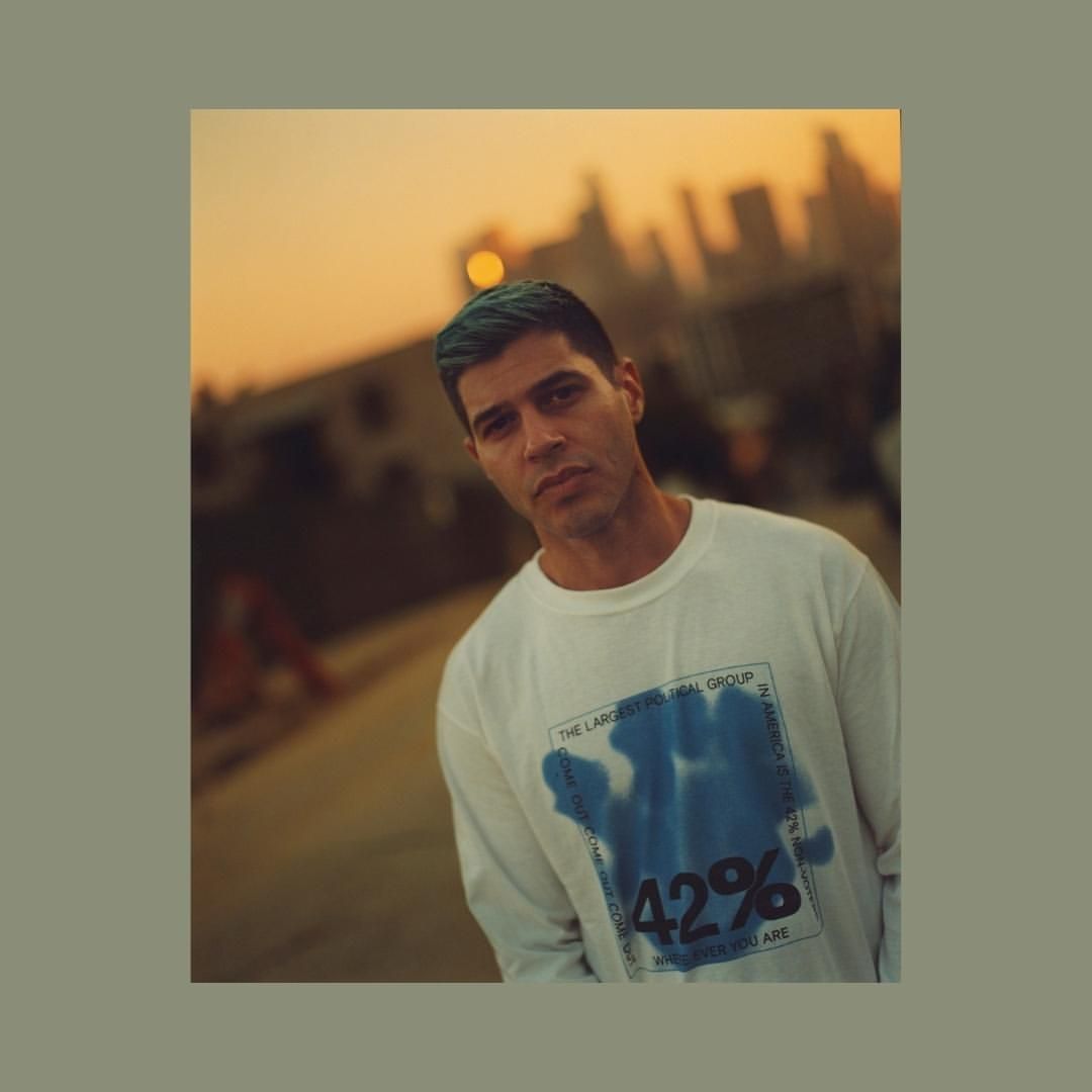Portrait of a young man with green-tinted hair, wearing a graphic sweatshirt, with a cityscape and golden sunset in the background.