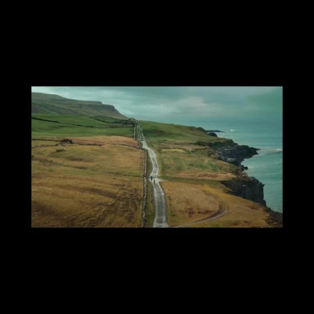 Scenic aerial view of two people walking on a rural road along a coastal hillside with green fields and the ocean in the background.