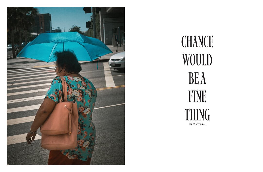 Woman with blue umbrella crossing city street, floral blouse, light brown purse; urban environment, crosswalk, sunny day.