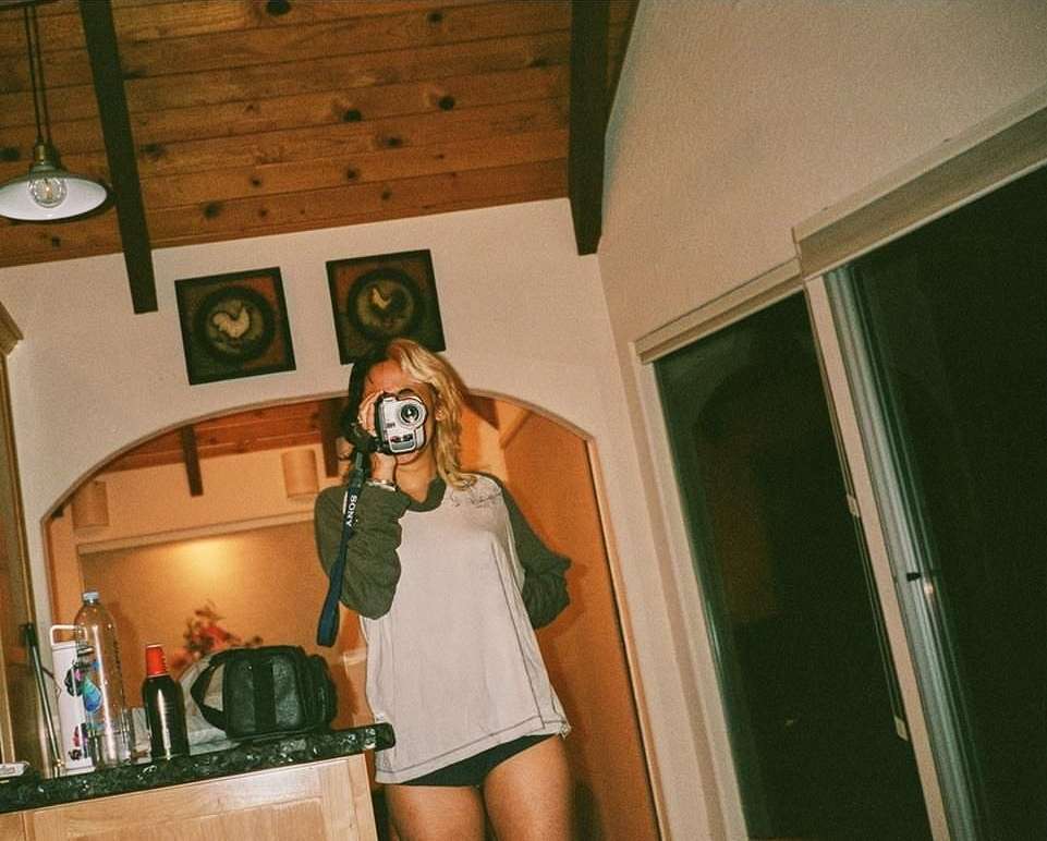 An adult female stands indoors, holding a camera up to her face, with a kitchen countertop and window visible in the background.