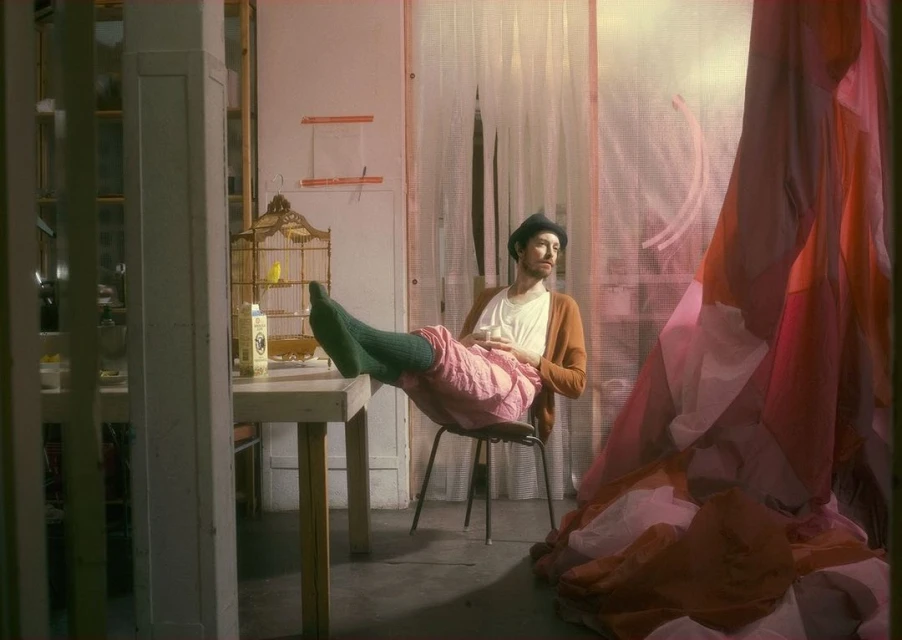 Indoor portrait of a man in a hat, cardigan, and pink pants, relaxing with his feet on a table, birdcage and a patchwork textile.