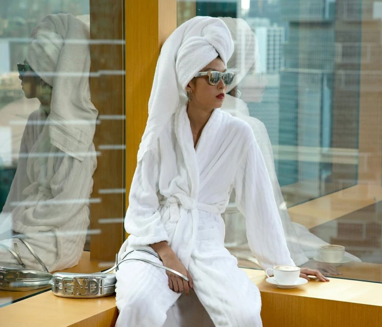 Woman in white bathrobe and head towel sits in front of a window with a cup of coffee, accessorized with sunglasses and purse.
