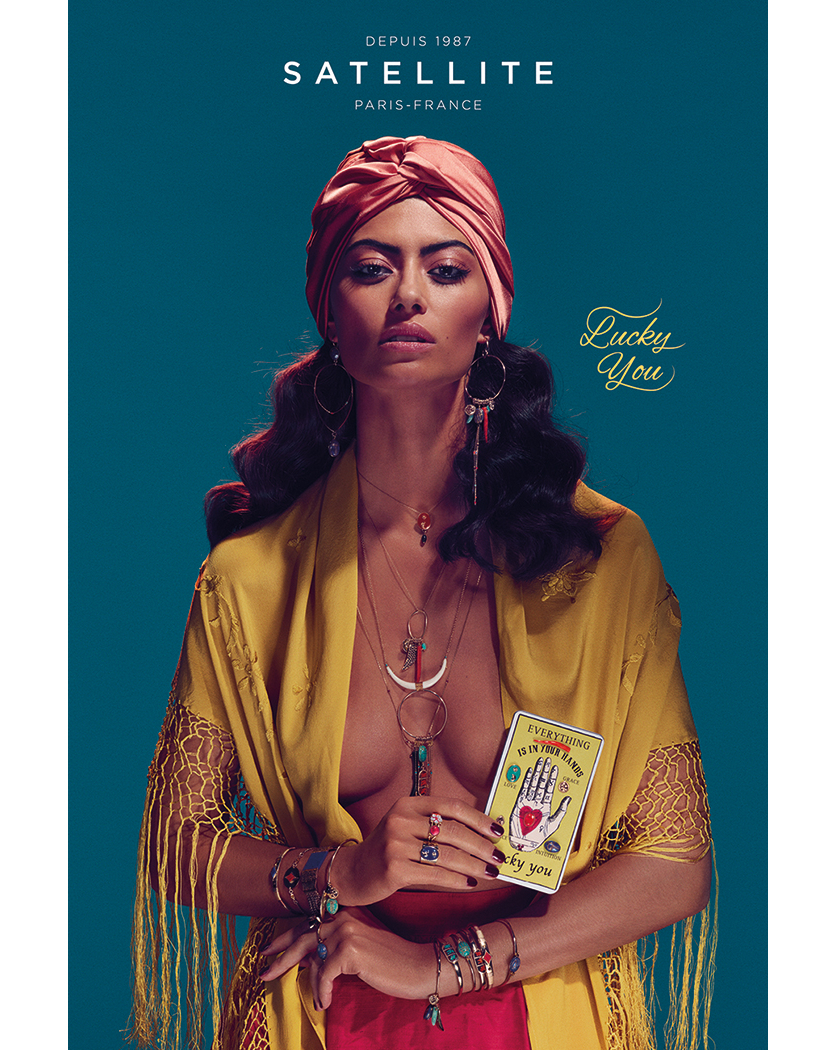 A woman in a headscarf and gold robe holds a palm reading card against a teal background in this fashion advertisement.