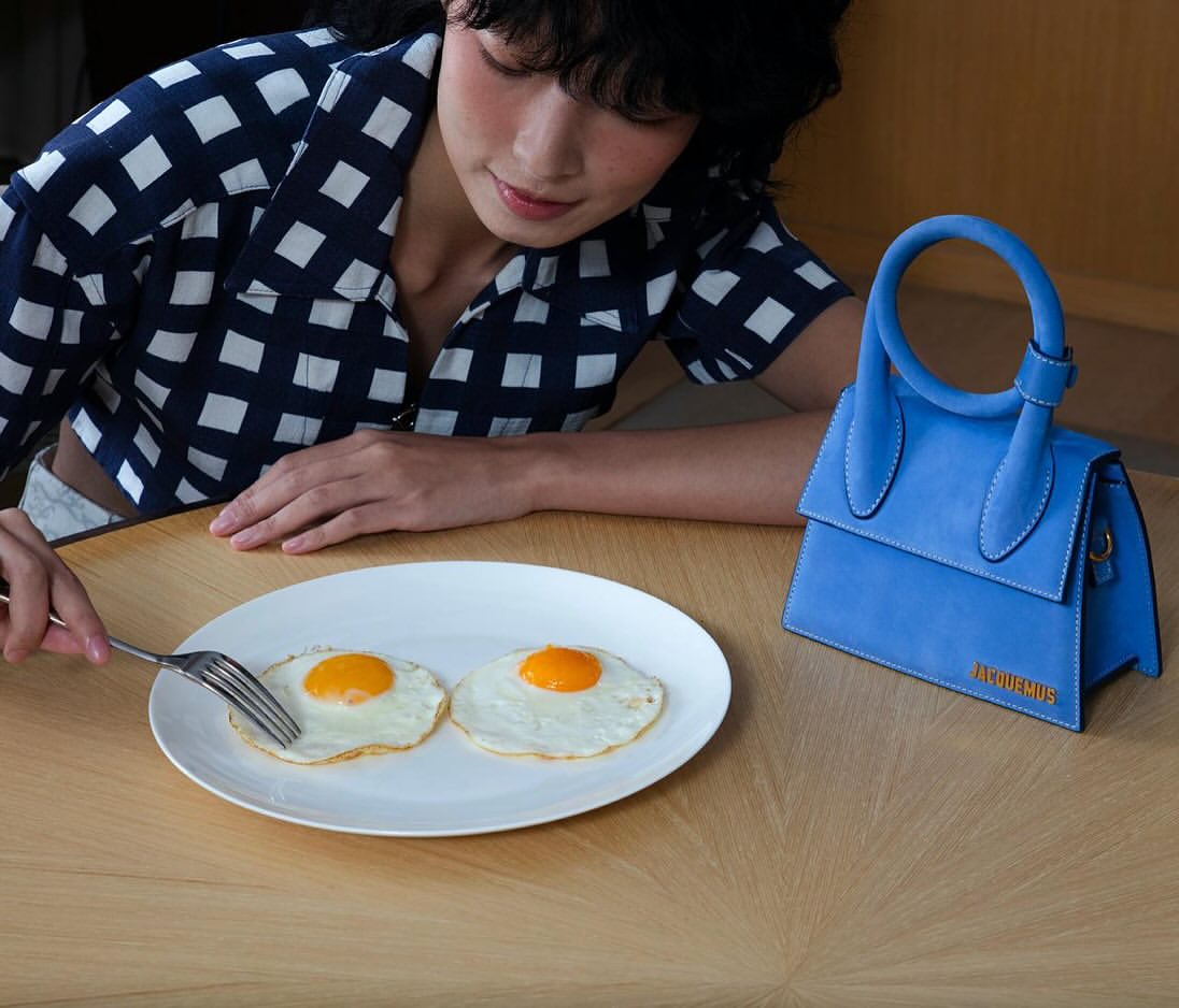 Woman in a patterned shirt at a table with fried eggs on a white plate and a small blue Jacquemus handbag; indoor setting.