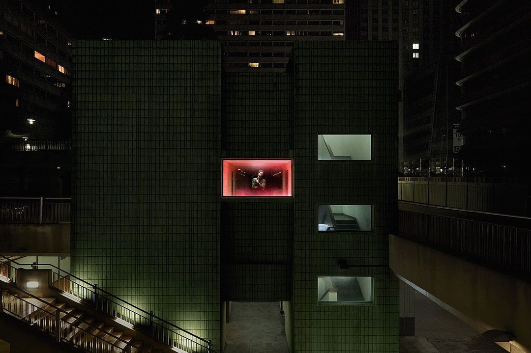 Night shot of a person in a brightly lit glass box on a green-tiled building, surrounded by urban architecture and dimly lit stairs.