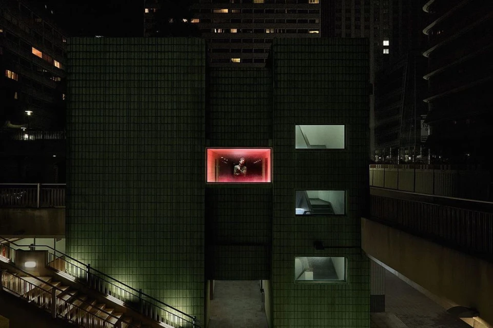 Night shot of a person in a brightly lit glass box on a green-tiled building, surrounded by urban architecture and dimly lit stairs.
