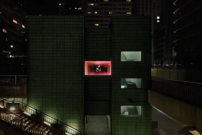 Night shot of a person in a brightly lit glass box on a green-tiled building, surrounded by urban architecture and dimly lit stairs.