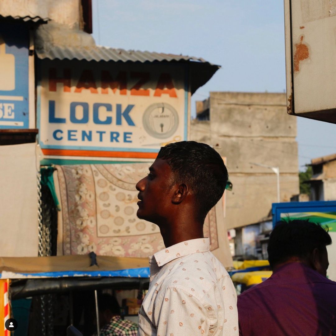 Young man in patterned shirt gazes upward outdoors in an urban setting with shop signs and neutral light in a medium-close shot.