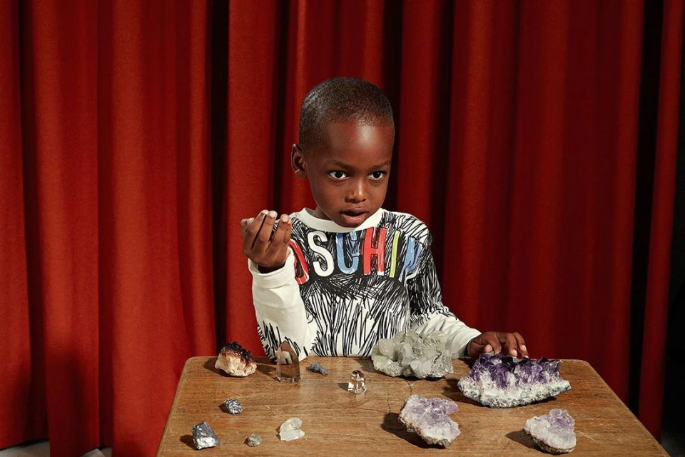 Young boy in a patterned shirt stands at a wooden table, displaying minerals and crystals against a backdrop of rich red curtains.