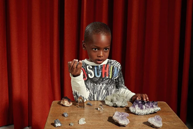 Young boy in a patterned shirt stands at a wooden table, displaying minerals and crystals against a backdrop of rich red curtains.