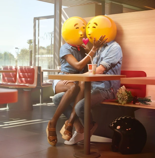 Two people with emoji heads at a restaurant, wearing casual denim outfits and sitting at a table with a drink, creating a playful and modern scene.