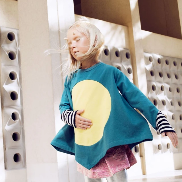 Child models teal and yellow long-sleeve tunic with striped sleeves, wearing a pink patterned skirt and silver leggings, inside a building.