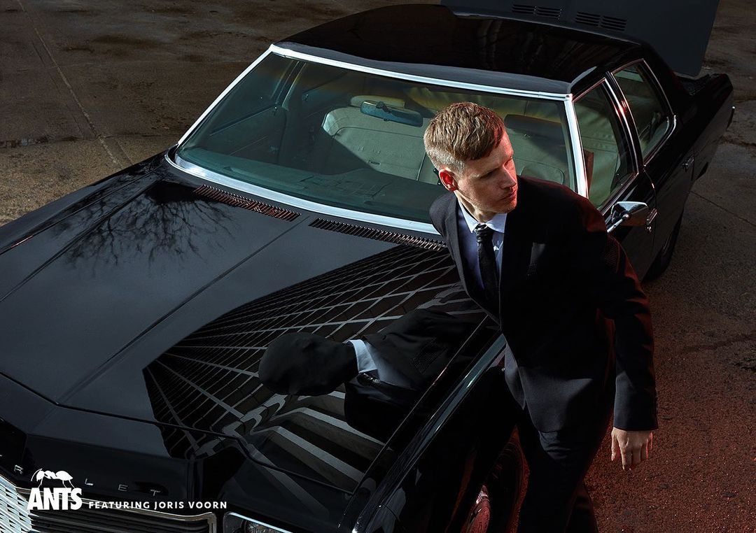 Stylish man in suit exits a black car with the trunk open, building reflection on the hood, evening outdoor setting.
