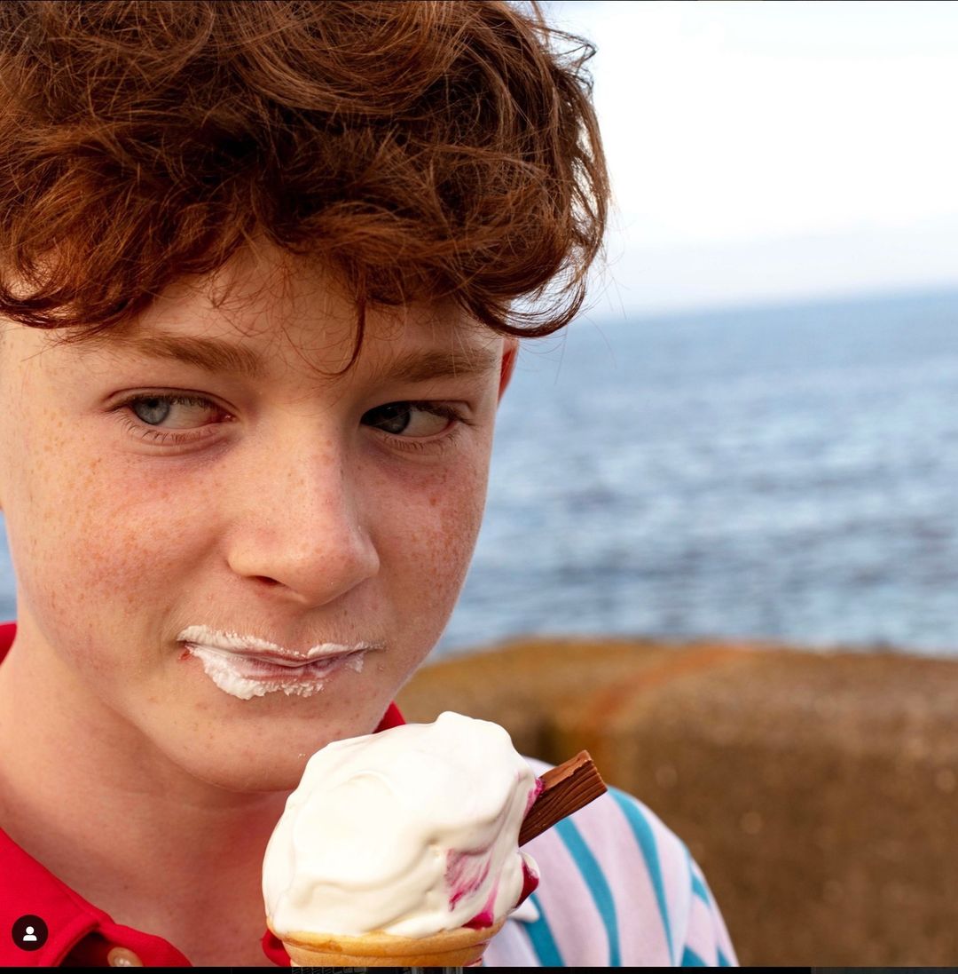 Boy with red hair eating ice cream cone with chocolate bar at beach; sea and horizon in background; summer food concept.