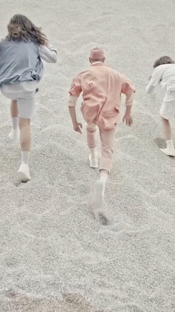 Three figures in pastel clothing run on a gray, gravel-like surface, their forms receding from the viewer in a stylized fashion shot.