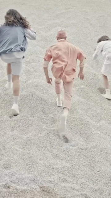 Three figures in pastel clothing run on a gray, gravel-like surface, their forms receding from the viewer in a stylized fashion shot.