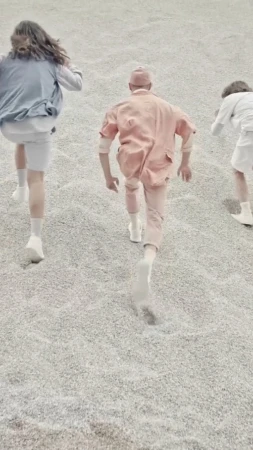 Three figures in pastel clothing run on a gray, gravel-like surface, their forms receding from the viewer in a stylized fashion shot.