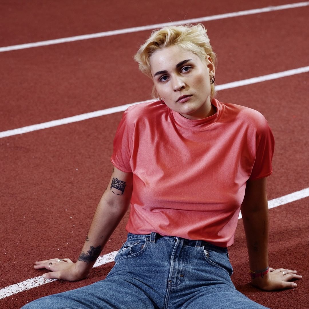 Woman with short blonde hair wearing a peach-colored shirt and jeans sits on a red running track with white lines.
