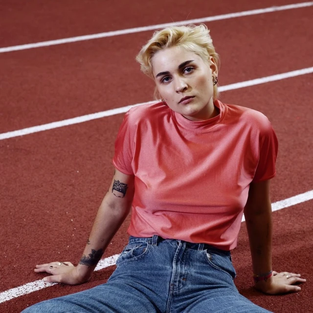 Woman with short blonde hair wearing a peach-colored shirt and jeans sits on a red running track with white lines.