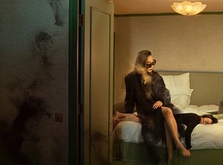 Two women in a dimly lit hotel room with one seated on the edge of the bed wearing a dark coat and sunglasses.