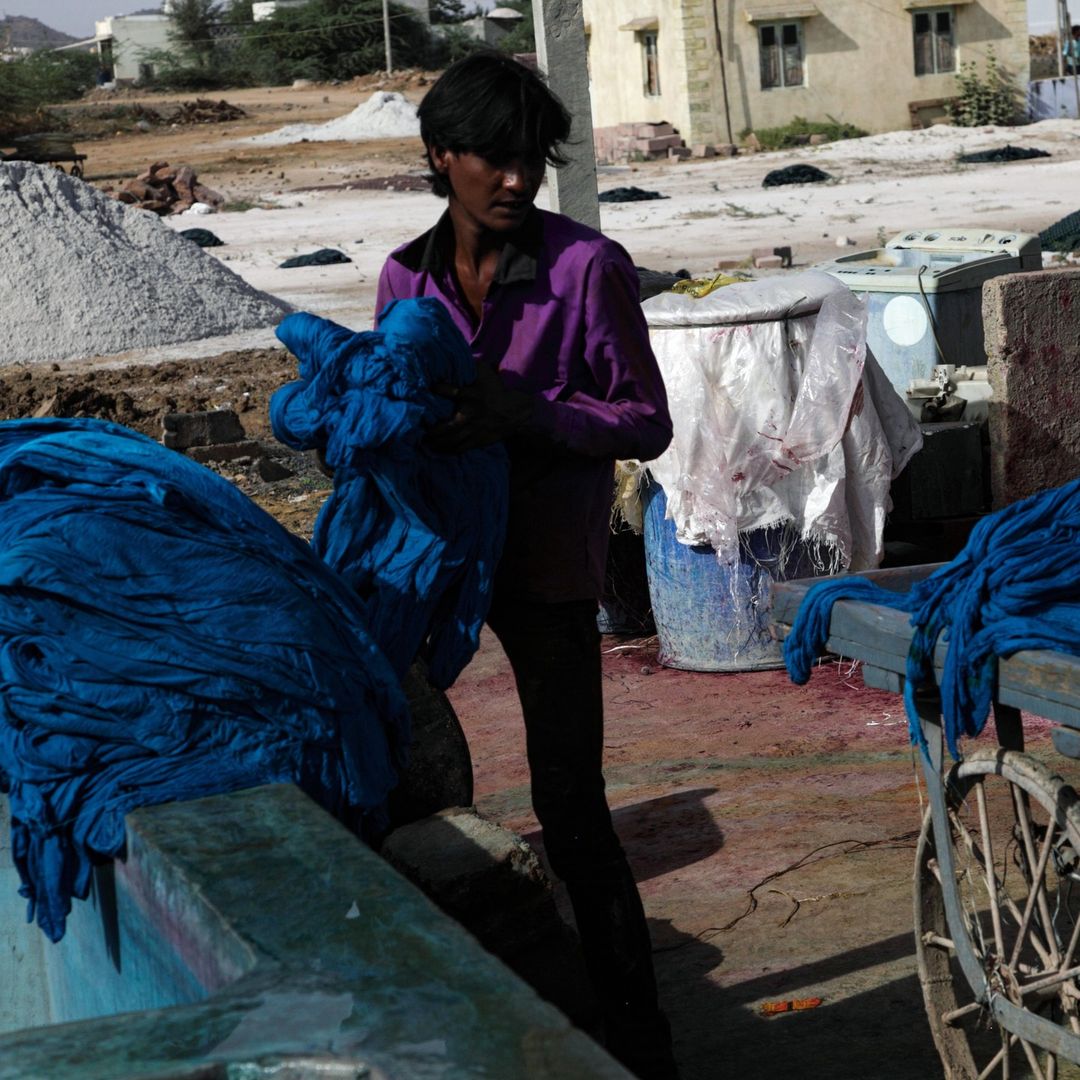 Person in a purple shirt handles draped blue textile outside near industrial containers and mounds of material, with a building in the background.