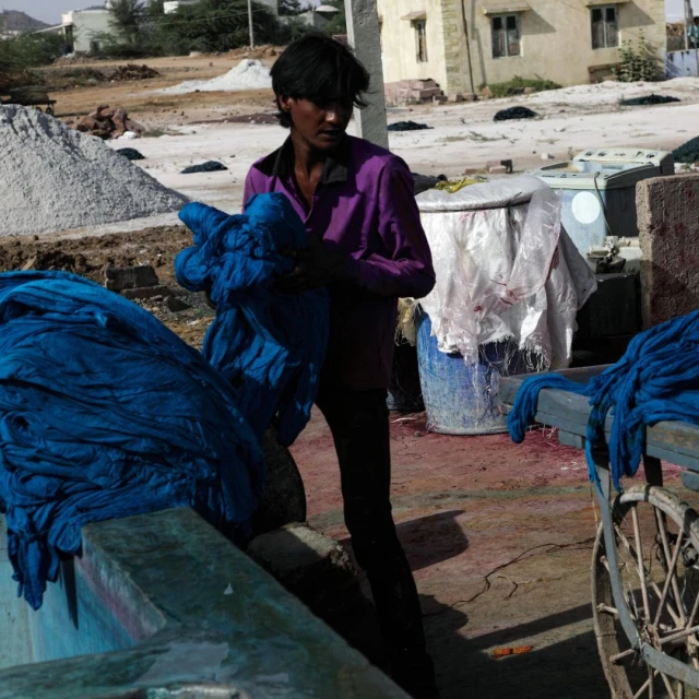 Person in a purple shirt handles draped blue textile outside near industrial containers and mounds of material, with a building in the background.