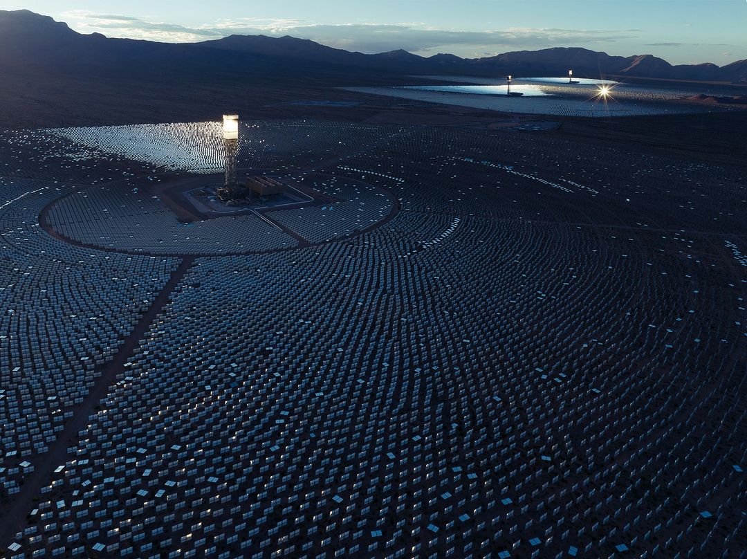Aerial view of a solar power plant with heliostat mirrors concentrating sunlight on a central receiver tower in the desert landscape.