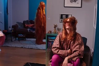 Indoor view of two adult females; one sits with her face covered by red hair, another is standing with full body covered in red hair.