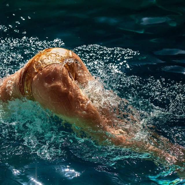 Woman swimming in a pool, underwater view with splashes and sunlight reflecting on the water's surface in an outdoor setting.